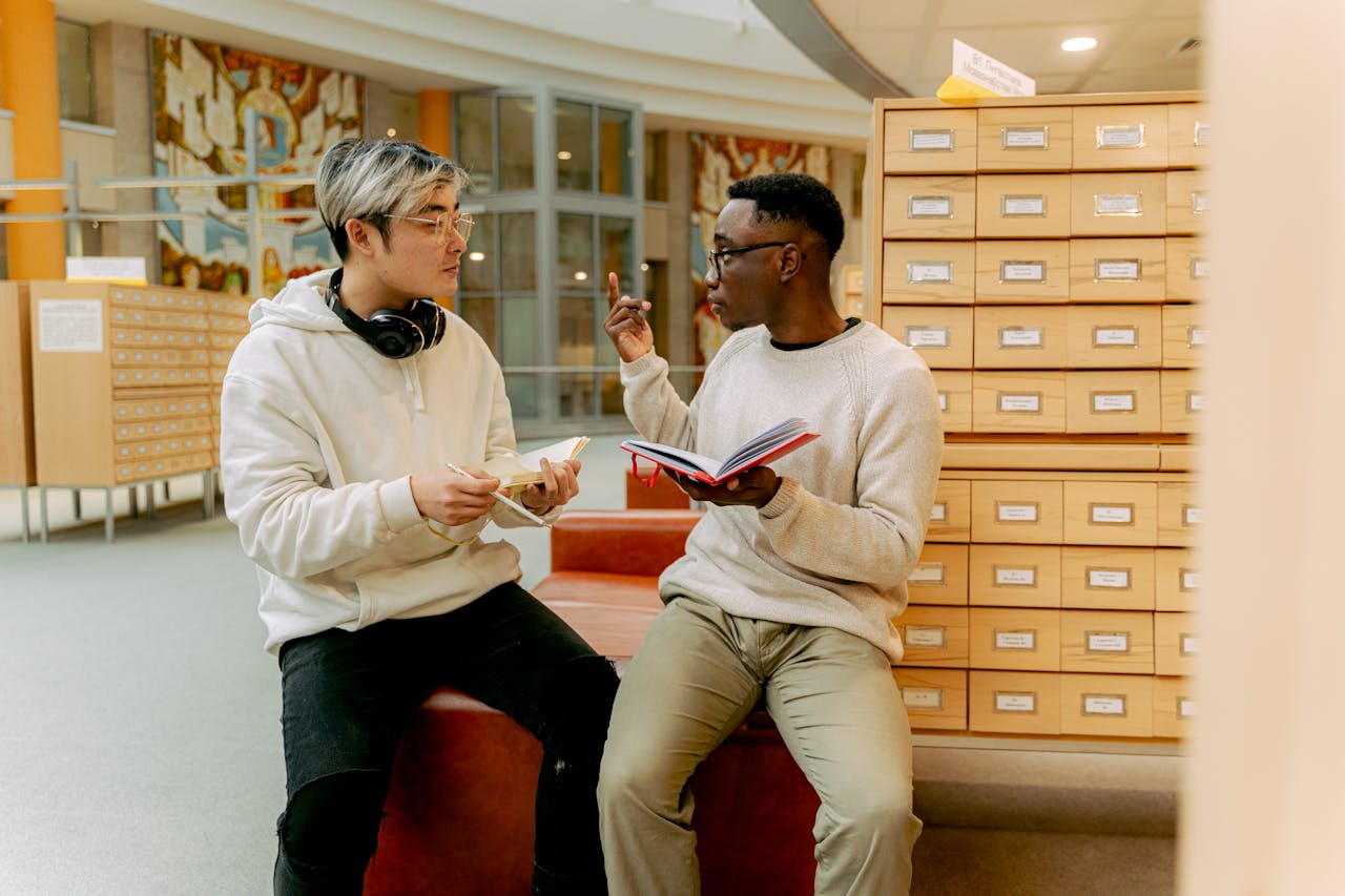 Two young men discussing in a library, exchanging ideas and holding notebooks.